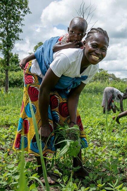A smiling farmer carrying her child on her back while working her field